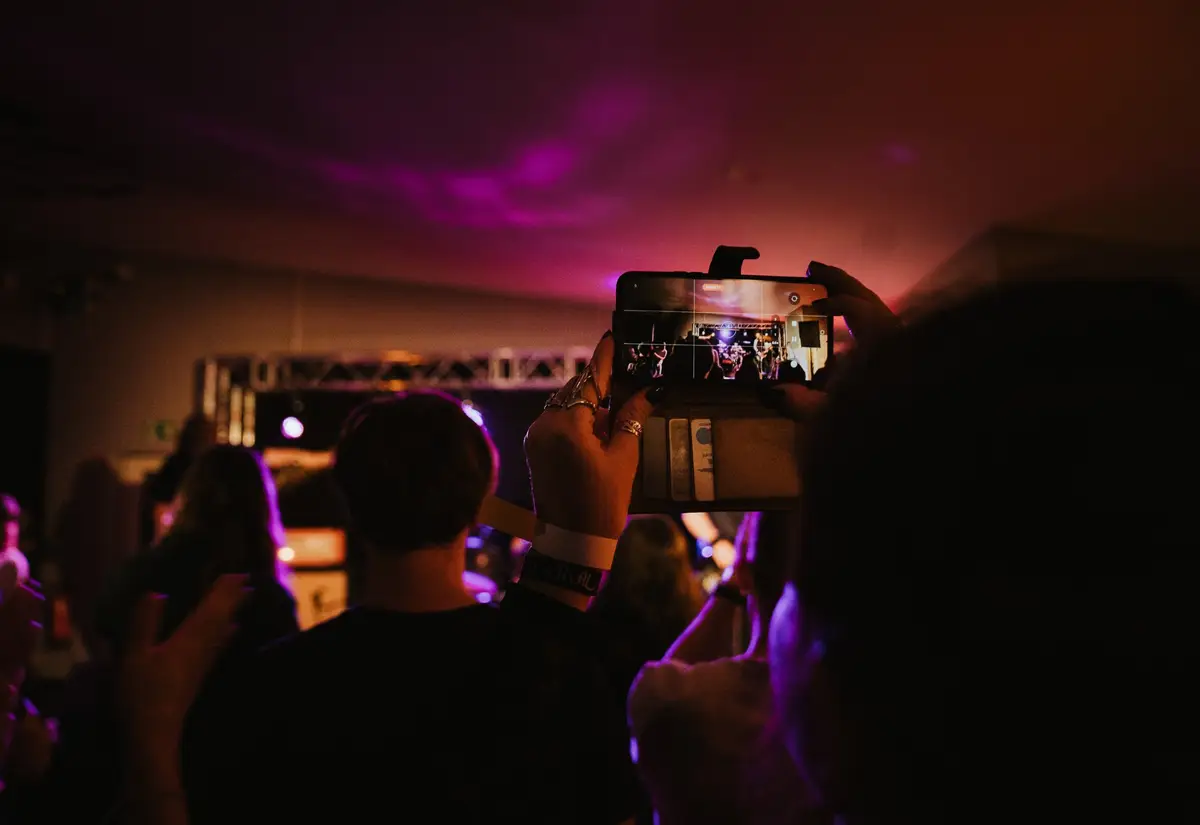 Konzertpublikum im KULT61, ein Smartphone filmt die Bühne im lila Licht